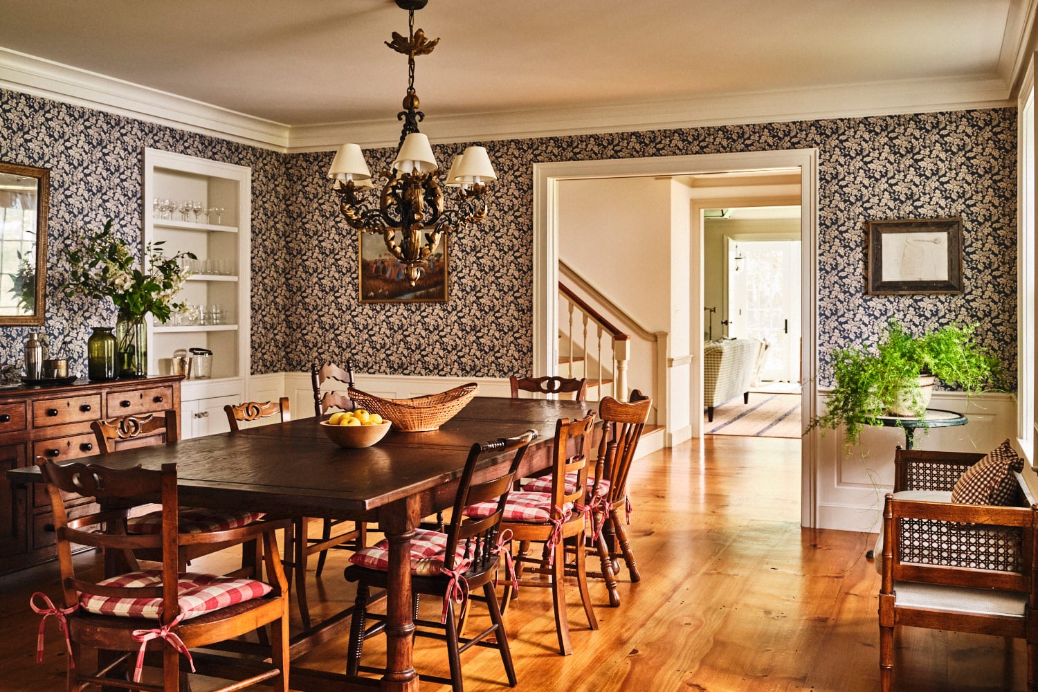 Dining room of main house at Vermont countryside compound Sarah Shetter designed for Hollywood writer-producers featuring Robert Kime oak-leaf-motif wallpaper, an early-19th-century French mirror hanging above a 19th-century English oak dresser, a Rococo style six-arm chandelier, an Scotish wood table and mismatched wood chairs and a faux bamboo and cane bench