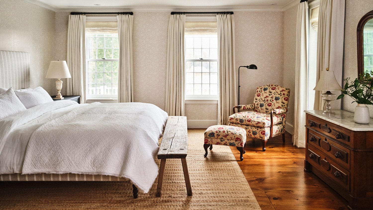 Primary bedroom of main house at Vermont countryside compound Sarah Shetter designed for Hollywood writer-producers featuring an antique armchair reupholstered in a Pierre Frey print and a ca. 1900 Victorian chest with marble top and an alabaster lamp from Liz's Antique Hardware on the nightstand. The bed is upholstered in ecru and white vertical stripes with white bedding.