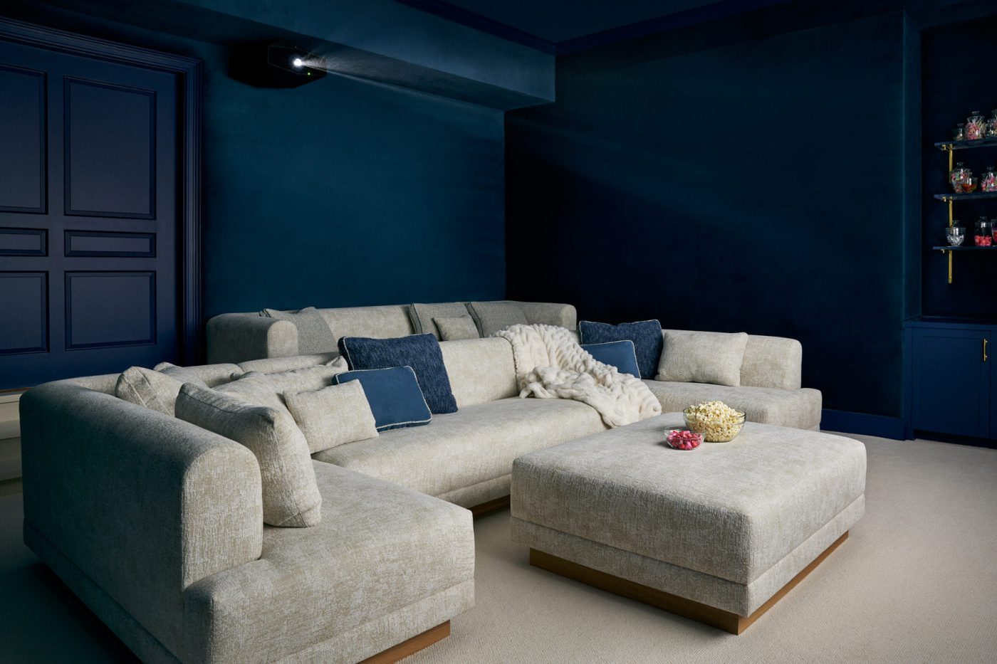 Media room with large off-white sectional and sofa and ottoman and deep blue walls and ceiling on lower level of Southampton house with interiors by Janine Carendi MacMurray or Area Interior Design