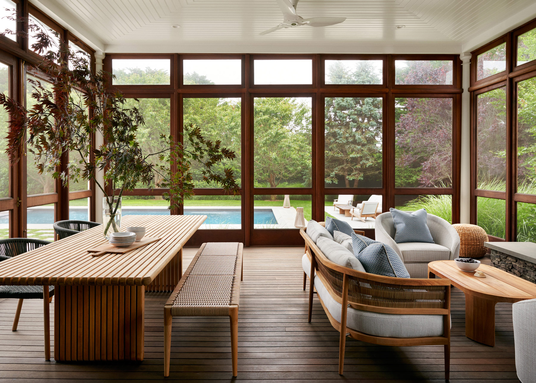 Teak furniture in dining area and sitting area of the screened-in porch of Southampton house with interiors by Janine Carendi MacMurray or Area Interior Design