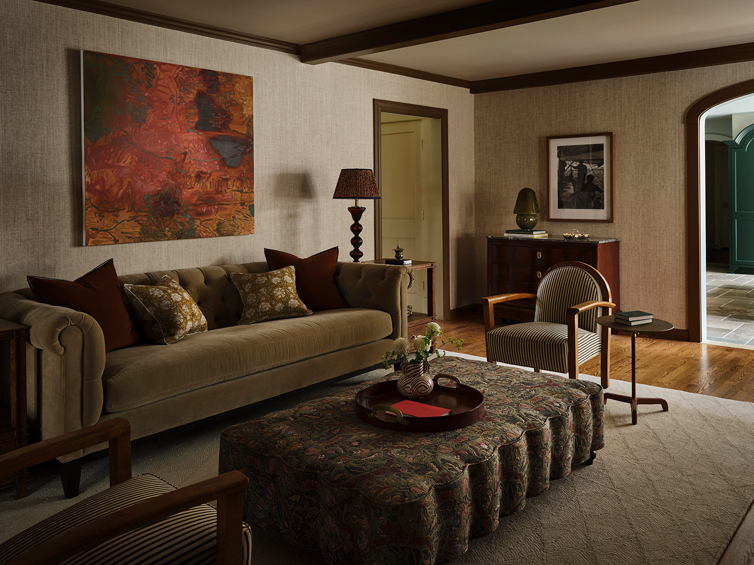 Phillip Jeffries neutral wallpaper, an Anne Griffith abstract painting, rollarm chesterfield-style velvet sofa, Art Deco–style armchairs, a scalloped ottoman covered in a Zak + Fox fabric and amber Venetian-glass table lamps from Antique Resourcesin the living room of a New Hampshire house with interiors by designer Zoe Feldman