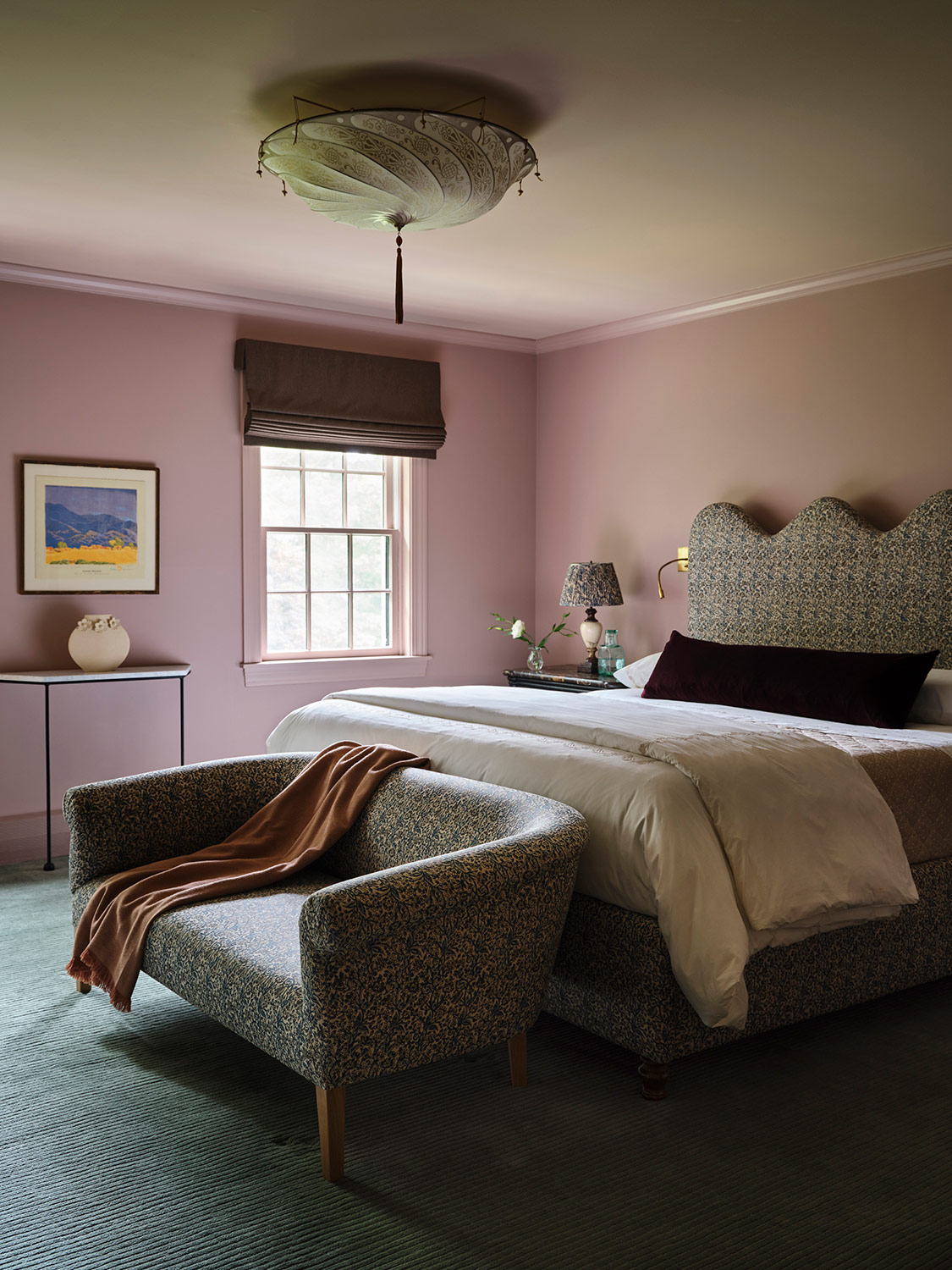 Primary bedrooms with rose-colored walls, green carpet and bed, end-of-bed settee and bedside lamp shades all upholstered in the same botanical print in a New Hampshire house with interiors by designer Zoe Feldman