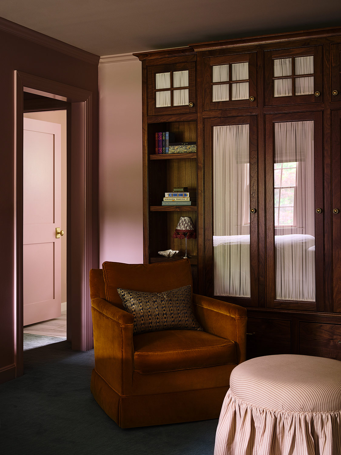 A skirted ottoman and an armchair recovered in a rusty-colored fabric, in front of a new wall of walnut-wood glass-fronted storage cabinets and shelves and drawers in the primary bedroom of a New Hampshire house with interiors by designer Zoe Feldman