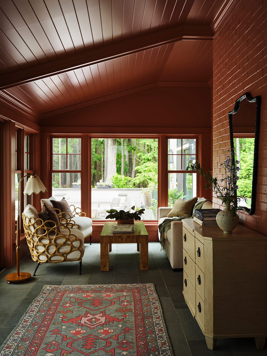 Cane armchairs, scalloped lampshade floor lamp. chest and tribal rug in the terracotta-colored sunroom of a New Hampshire house with interiors by designer Zoe Feldman