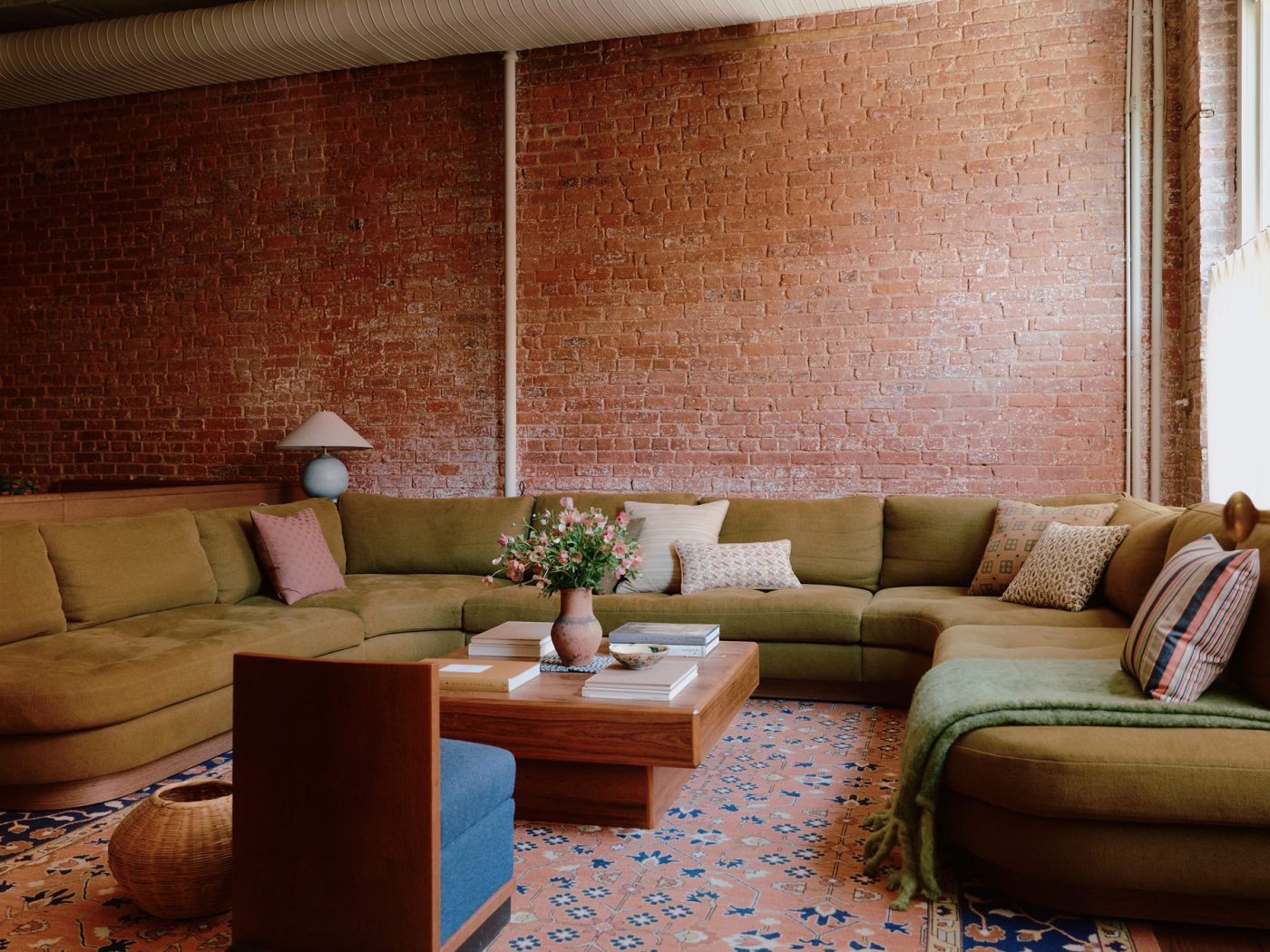 Large olive-green u-shaped built-in sectional sofa in front of large windows atop a salmon and blue oriental carpet with a vintage chair and coffee table, in the living area of a New York loft designed by Sarah Zames and Colin Stief, of the Brooklyn-based studio General Assembly