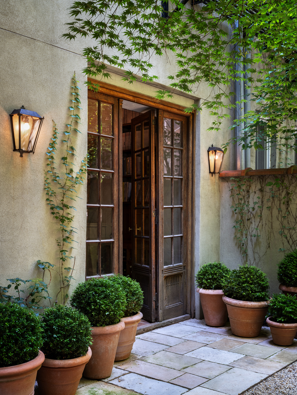 Residential courtyard garden in Manhattan's West Village designed by the Brooklyn studio Harrison Green and incorporating containers planted with boxwood dotting a stone patio in front of a wooden-framed glass door and sidelights
