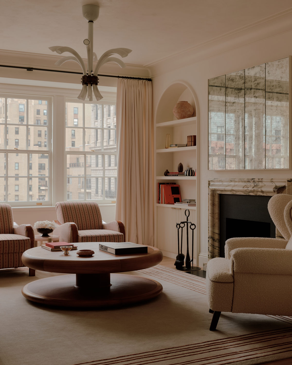 Living room with Swedish biedermeier arm chairs, leather-topped round coffee table, and boucle-covered wingback chair in front of a marble mantel under antiqued mirrors beside built-in shelves in a duplex apartment on New York's Park Avenue designer by Bryan O'Sullivan