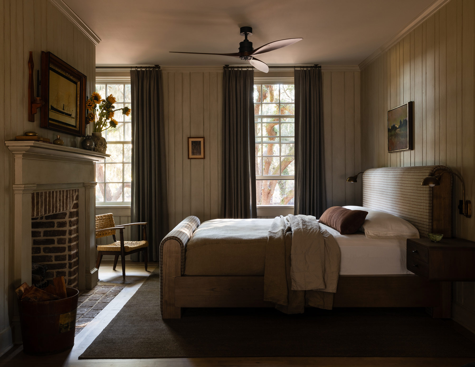 Primary bedroom with wood mantel and brick fireplace and upholstered bed and wood-paneled walls in a former agrarian building on South Carolina's Edisto Island reimagined as a vacation home by interior designer Sean Anderson and architect Will Wingfield