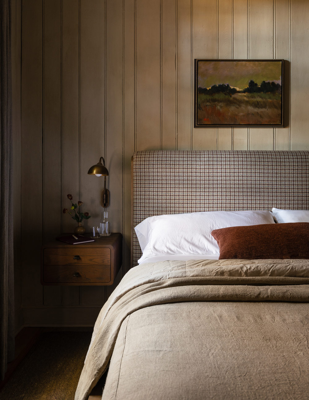 Primary bedroom with wood mantel and brick fireplace and upholstered bed and wood-paneled walls in a former agrarian building on South Carolina's Edisto Island reimagined as a vacation home by interior designer Sean Anderson and architect Will Wingfield