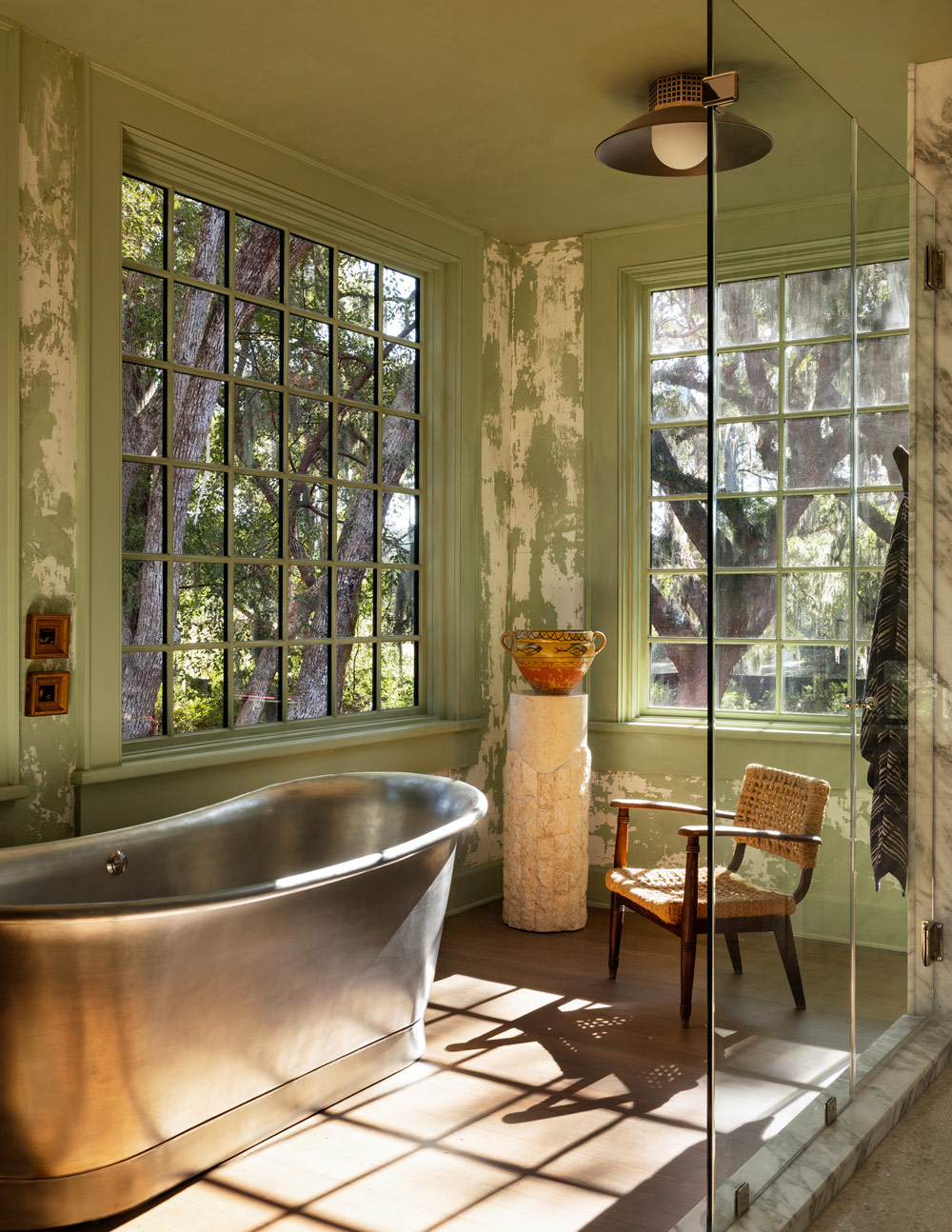 Primary bathroom with corner windows, metal soaking tube and glass enclosed shower, plus woven-string modern chair and urn on a plaster pedestal in a former agrarian building on South Carolina's Edisto Island reimagined as a vacation home by interior designer Sean Anderson and architect Will Wingfield