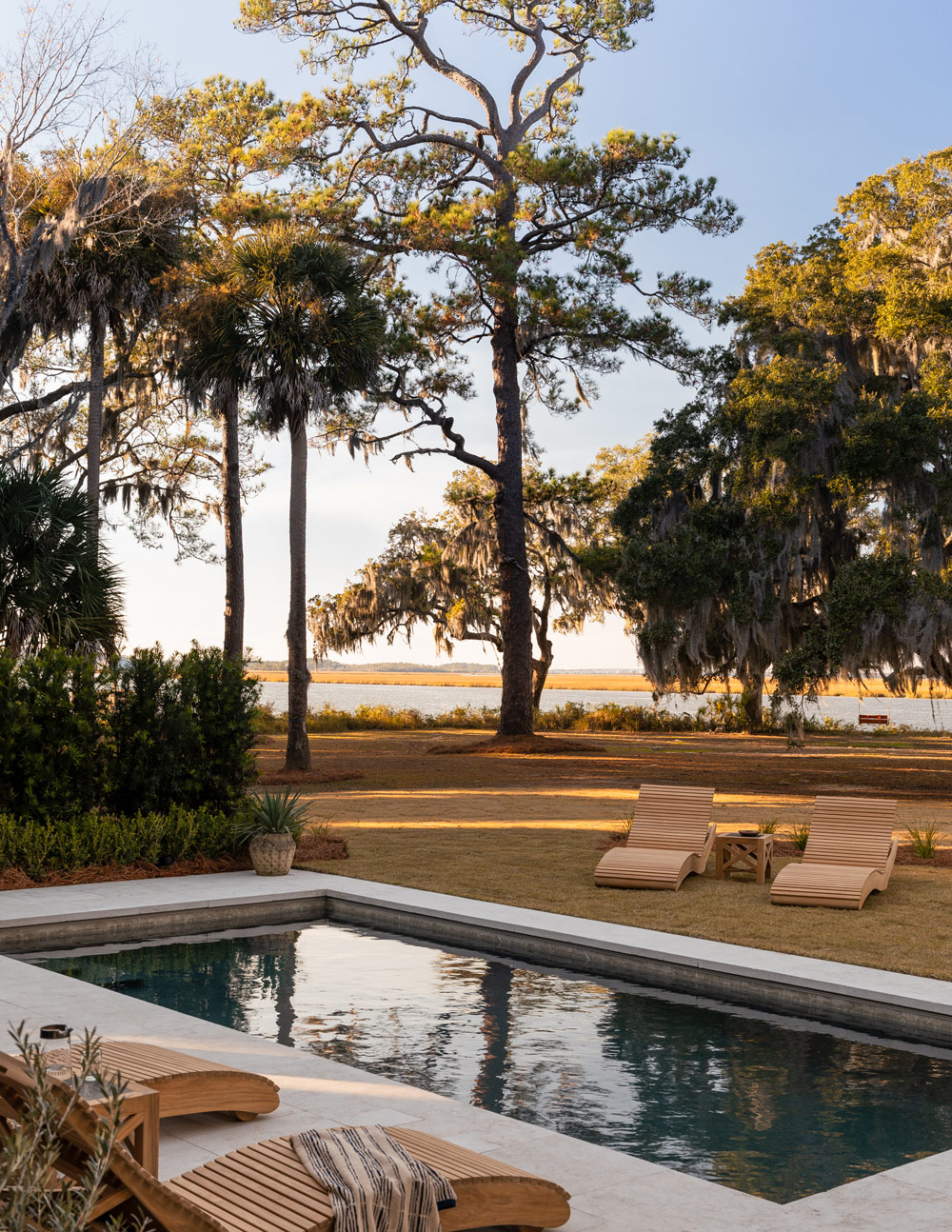 Pool area with teak chaises and views of South Edisto River of a property that's a former agrarian building on South Carolina's Edisto Island reimagined as a vacation home by interior designer Sean Anderson and architect Will Wingfield, Landscape designed by Glen R. Gardner