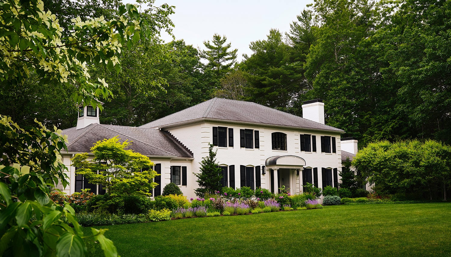 Exterior of a neoclassically inspired white stucco New Hampshire house with a black roof, windows and shutters and corner coping and, inside, new interiors by Zoe Feldman