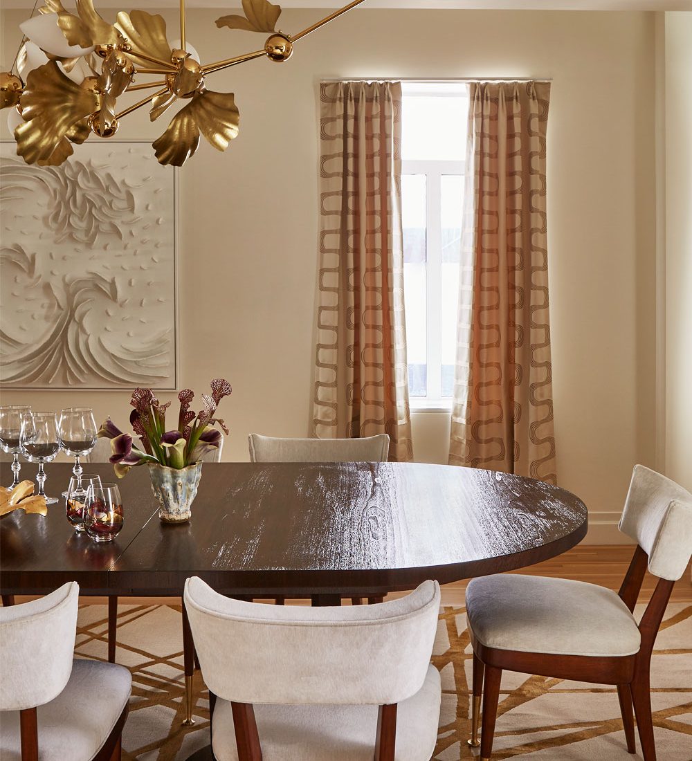 A gold-and-white lotus flower chandelier by Rosie Li hung over a brown wood oval table surrounded by brown wood chairs with off-white seats and back in the dining room of a penthouse apartment on New York City's Park Avenue designed by Young Huh